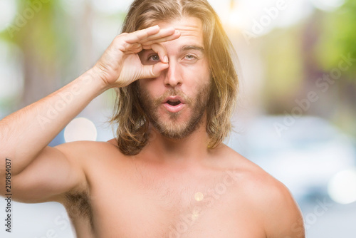 Young handsome shirtless man with long hair showing sexy body over isolated background doing ok gesture shocked with surprised face, eye looking through fingers. Unbelieving expression.