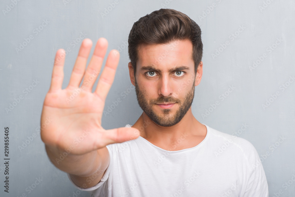 Handsome young man over grey grunge wall with open hand doing stop sign with serious and confident expression, defense gesture - obrazy, fototapety, plakaty 