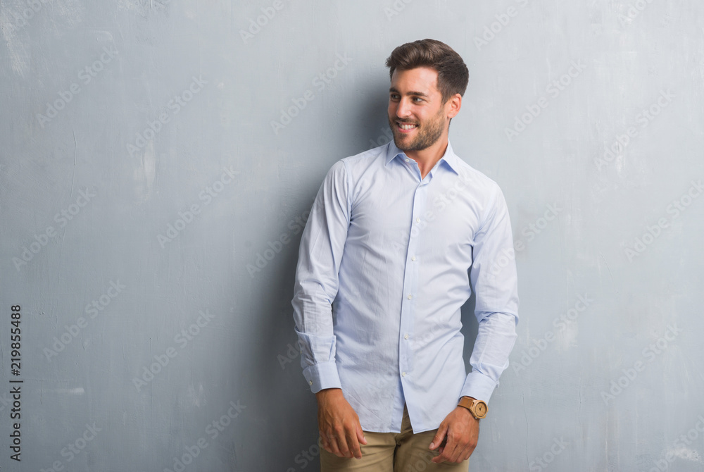 Handsome young business man over grey grunge wall wearing elegant shirt looking away to side with smile on face, natural expression. Laughing confident.