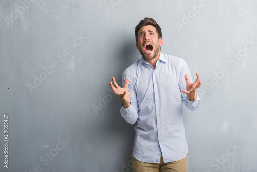 Handsome young business man over grey grunge wall wearing elegant shirt crazy and mad shouting and yelling with aggressive expression and arms raised. Frustration concept. © Krakenimages.com