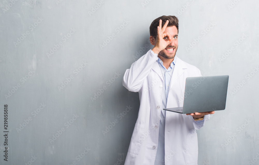 Handsome young man over grey grunge wall wearing white coat using laptop with happy face smiling doing ok sign with hand on eye looking through fingers Handsome young man over grey grunge wall wearing white coat using laptop with happy face smiling doing ok sign with hand on eye looking through fingers