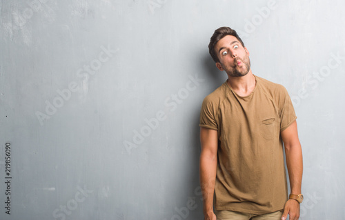 Handsome young man over grey grunge wall making fish face with lips, crazy and comical gesture. Funny expression. © Krakenimages.com