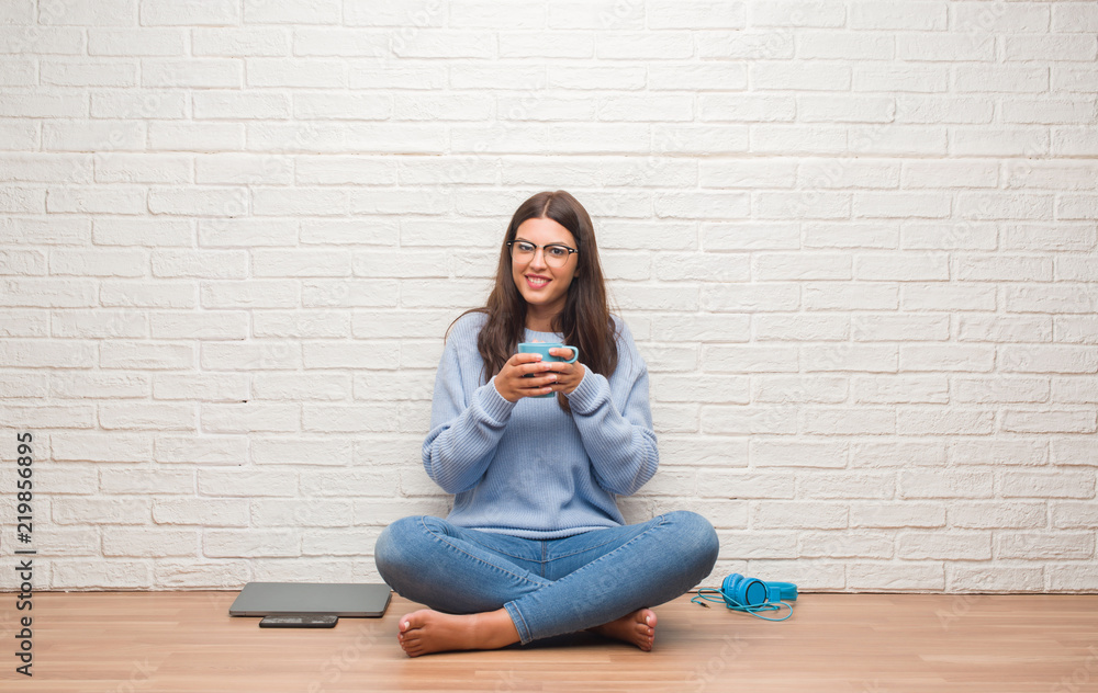 Young brunette woman sitting on the floor over white brick wall drinking coffee with a happy face standing and smiling with a confident smile showing teeth Young brunette woman sitting on the floor over white brick wall drinking coffee with a happy face standing and smiling with a confident smile showing teeth