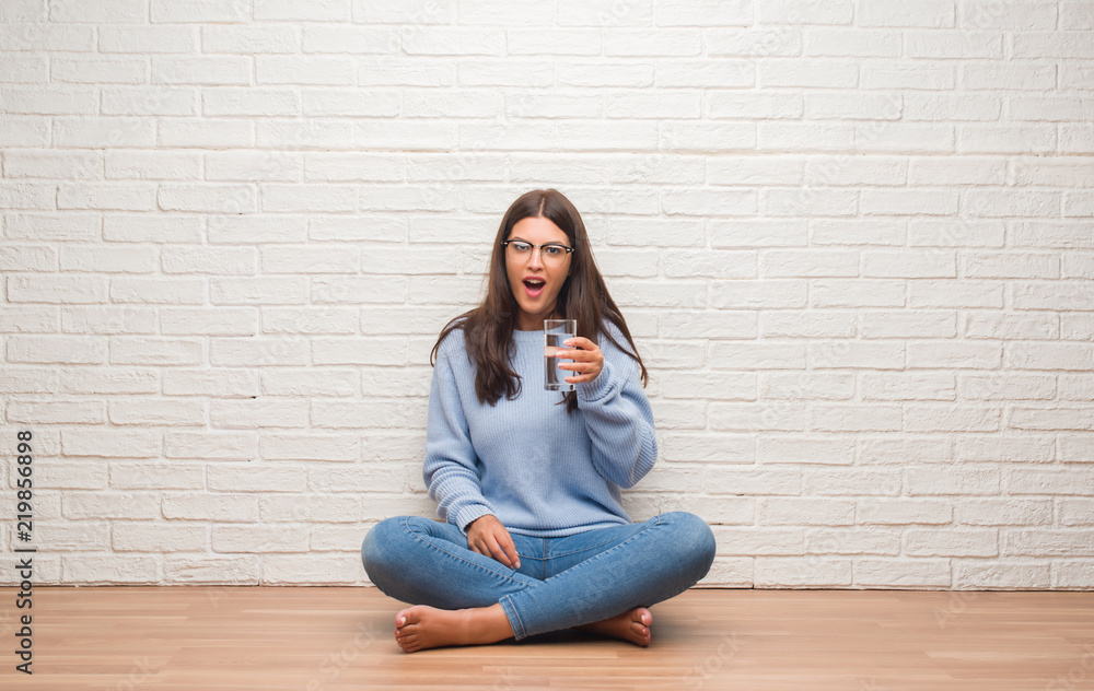 Young brunette woman sitting on the floor drinking glass of water scared in shock with a surprise face, afraid and excited with fear expression Young brunette woman sitting on the floor drinking glass of water scared in shock with a surprise face, afraid and excited with fear expression