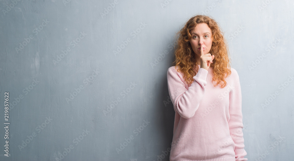 Young redhead woman over grey grunge wall wearing pink sweater asking to be quiet with finger on lips. Silence and secret concept. Young redhead woman over grey grunge wall wearing pink sweater asking to be quiet with finger on lips. Silence and secret concept.