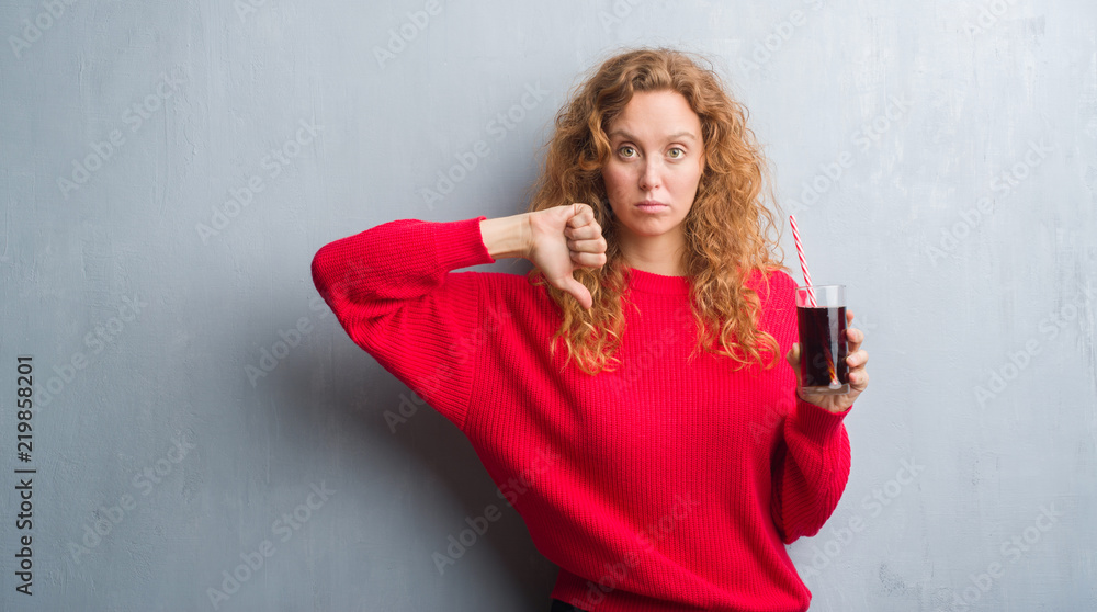 Young redhead woman over grey grunge wall drinking soda refreshment with angry face, negative sign showing dislike with thumbs down, rejection concept Young redhead woman over grey grunge wall drinking soda refreshment with angry face, negative sign showing dislike with thumbs down, rejection concept