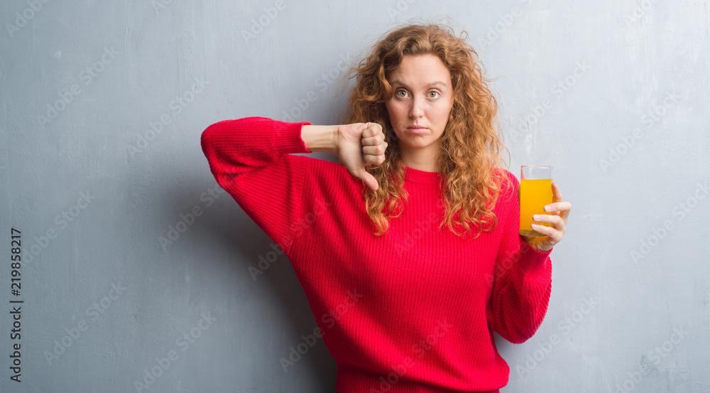 Young redhead woman over grey grunge wall drinking a glass of orange juice with angry face, negative sign showing dislike with thumbs down, rejection concept Young redhead woman over grey grunge wall drinking a glass of orange juice with angry face, negative sign showing dislike with thumbs down, rejection concept