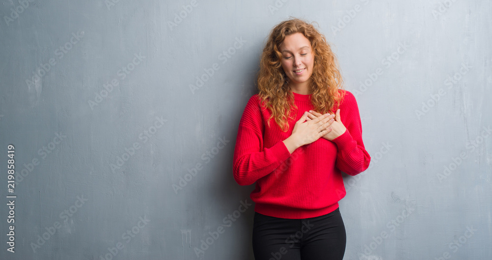 Young redhead woman over grey grunge wall wearing red sweater smiling with hands on chest with closed eyes and grateful gesture on face. Health concept.