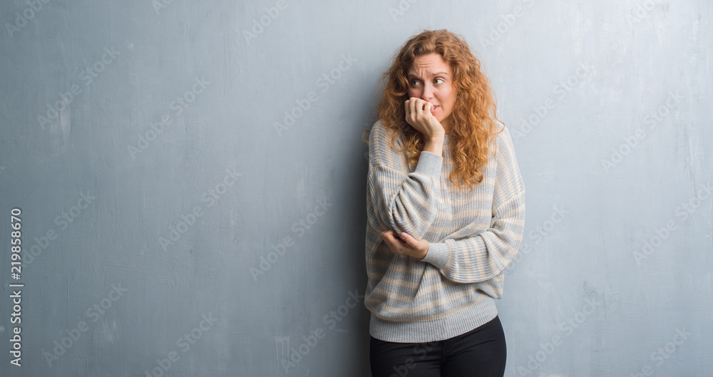 Fototapeta premium Young redhead woman over grey grunge wall looking stressed and nervous with hands on mouth biting nails. Anxiety problem.