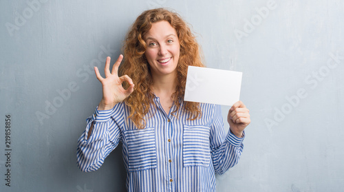 Young redhead woman over grey grunge wall holding blank card doing ok sign with fingers, excellent symbol