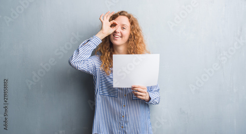 Young redhead woman over grey grunge wall holding blank paper sheet with happy face smiling doing ok sign with hand on eye looking through fingers