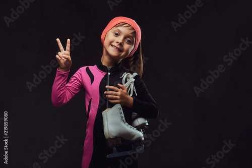 Happy little skater girl dressed in sportswear holds ice skates on a shoulder and shows victory hand. Isolated on dark textured background. © Fxquadro