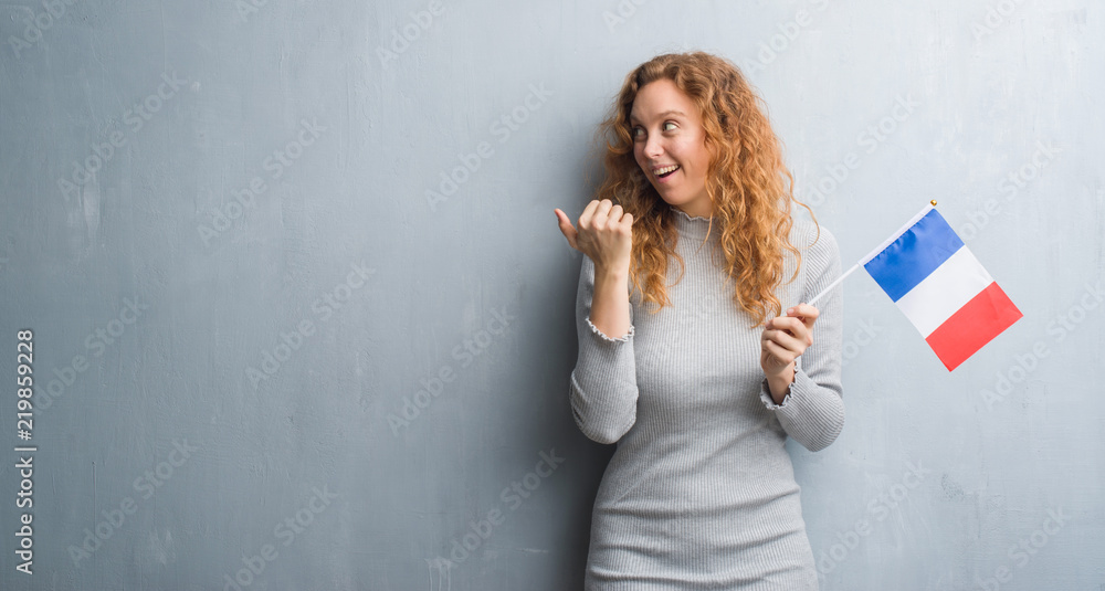 Young redhead woman over grey grunge wall holding flag of France pointing and showing with thumb up to the side with happy face smiling Young redhead woman over grey grunge wall holding flag of France pointing and showing with thumb up to the side with happy face smiling