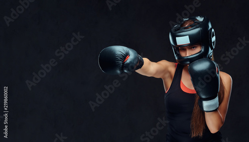 Sportive woman in sportswear wearing a protective helmet and boxing gloves, training in gym. Isolated on a dark textured background. © Fxquadro