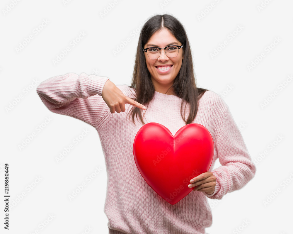 Young hispanic woman holding heart in love with surprise face pointing finger to himself Young hispanic woman holding heart in love with surprise face pointing finger to himself