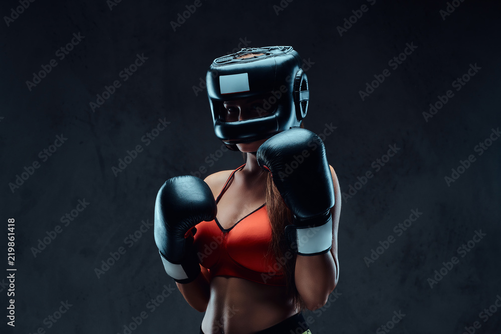 Sportive woman in sports bra wearing a protective helmet and boxing gloves,  posing in a studio. Isolated on a dark textured background. Stock Photo
