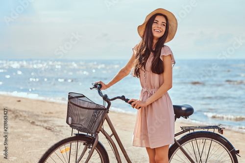 Laughing sensual girl with brown hair dressed in dress and hat posing with a bicycle on the beach on a sunny day.
