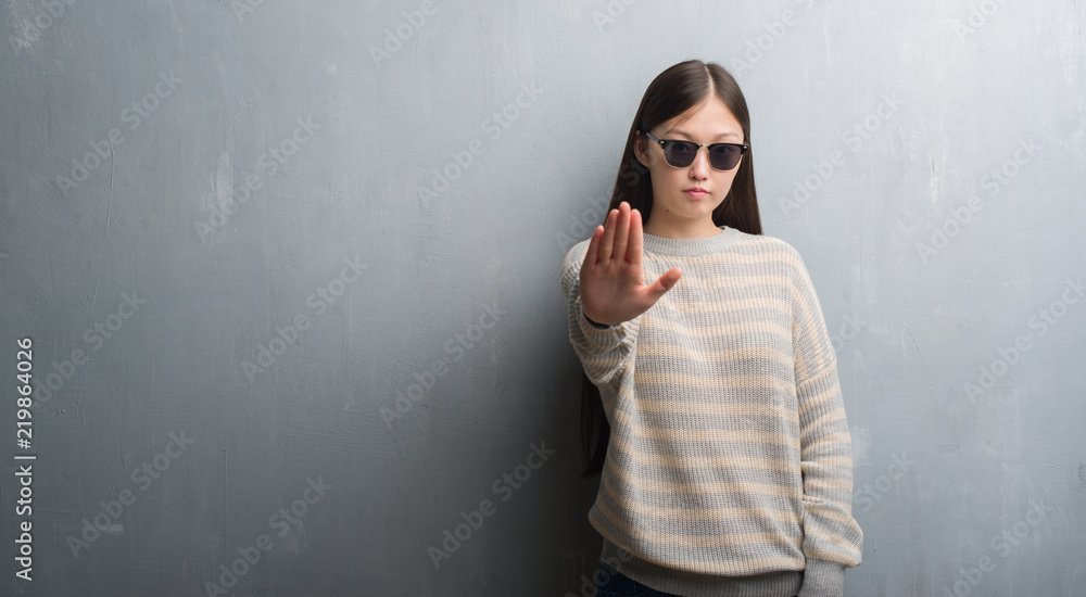 Young Chinese woman over grey wall wearing sunglasses with open hand doing stop sign with serious and confident expression, defense gesture Young Chinese woman over grey wall wearing sunglasses with open hand doing stop sign with serious and confident expression, defense gesture