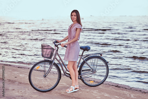 Smiling charming girl wearing dress walks with her bicycle on the beach.