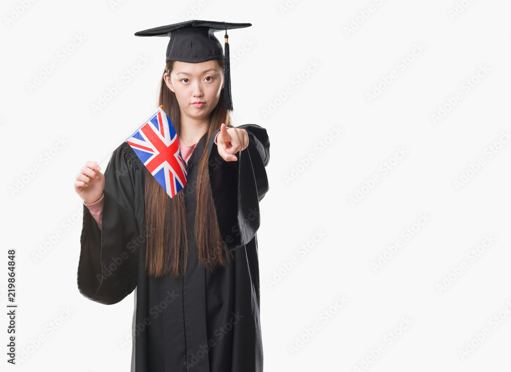 Young Chinese woman wearing graduate uniform holding United kingdom flag pointing with finger to the camera and to you, hand sign, positive and confident gesture from the front Young Chinese woman wearing graduate uniform holding United kingdom flag pointing with finger to the camera and to you, hand sign, positive and confident gesture from the front
