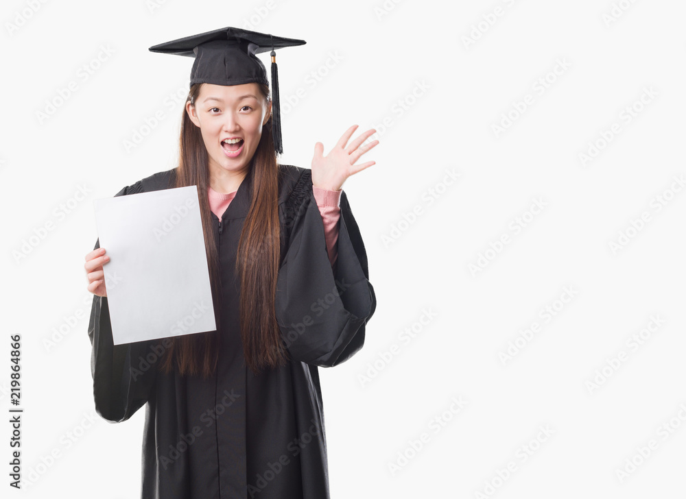 Young Chinese woman wearing graduate uniform holding paper degree very happy and excited, winner expression celebrating victory screaming with big smile and raised hands Young Chinese woman wearing graduate uniform holding paper degree very happy and excited, winner expression celebrating victory screaming with big smile and raised hands