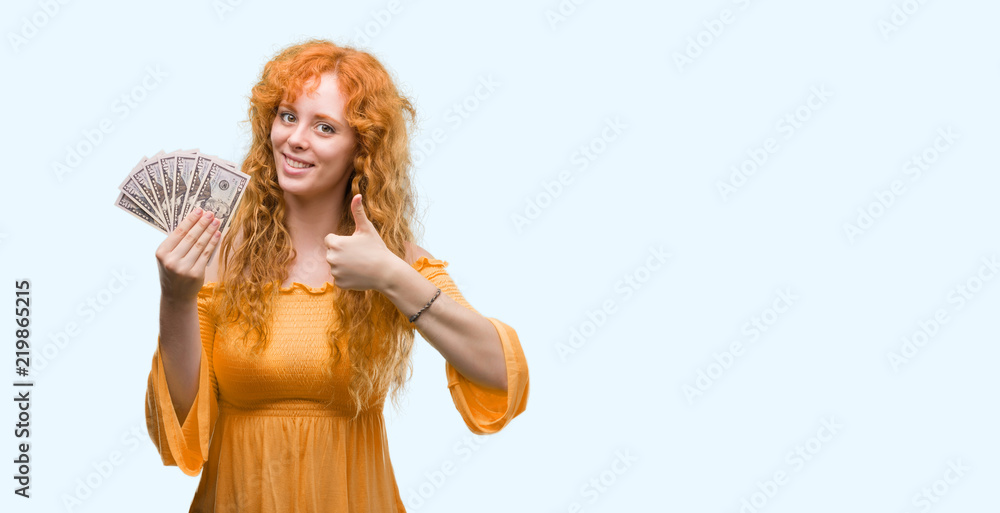 Young redhead woman holding dollars happy with big smile doing ok sign, thumb up with fingers, excellent sign Young redhead woman holding dollars happy with big smile doing ok sign, thumb up with fingers, excellent sign