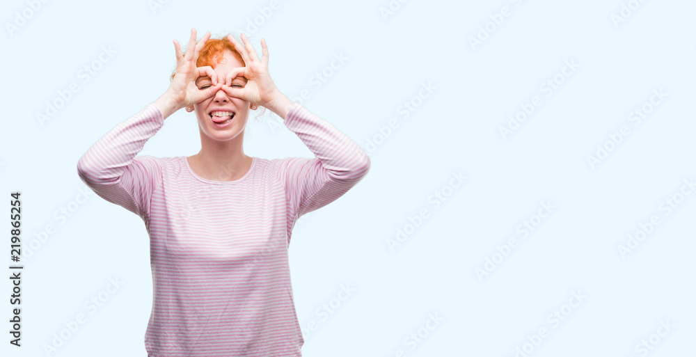 Young redhead woman doing ok gesture like binoculars sticking tongue out, eyes looking through fingers. Crazy expression. Young redhead woman doing ok gesture like binoculars sticking tongue out, eyes looking through fingers. Crazy expression.