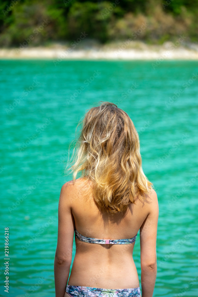 une jeune fille blonde, de dos, en maillot de bain  face à un lac au eaux vertes