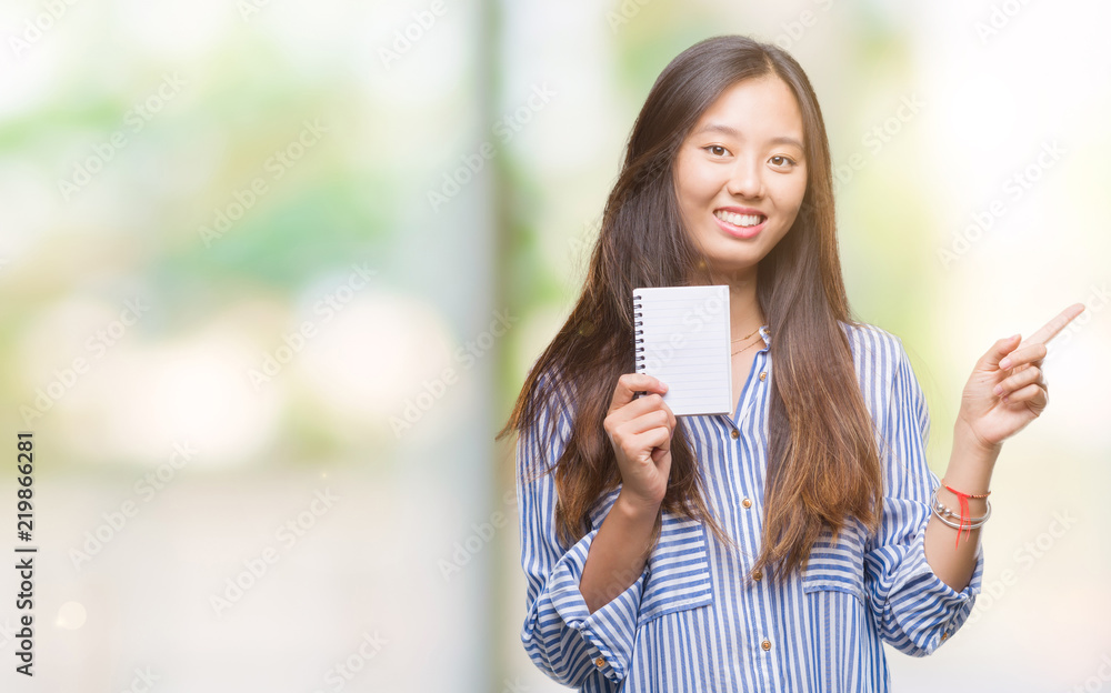 Young asian woman holding notebook over isolated background very happy pointing with hand and finger to the side