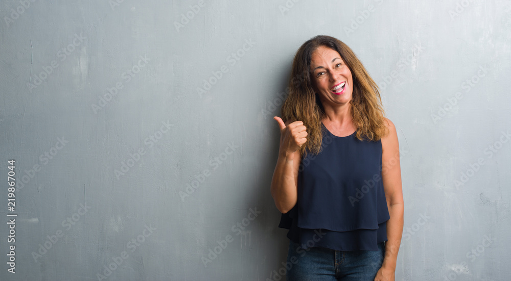 Fototapeta premium Middle age hispanic woman standing over grey grunge wall smiling with happy face looking and pointing to the side with thumb up.