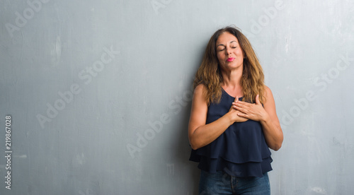 Middle age hispanic woman standing over grey grunge wall smiling with hands on chest with closed eyes and grateful gesture on face. Health concept.