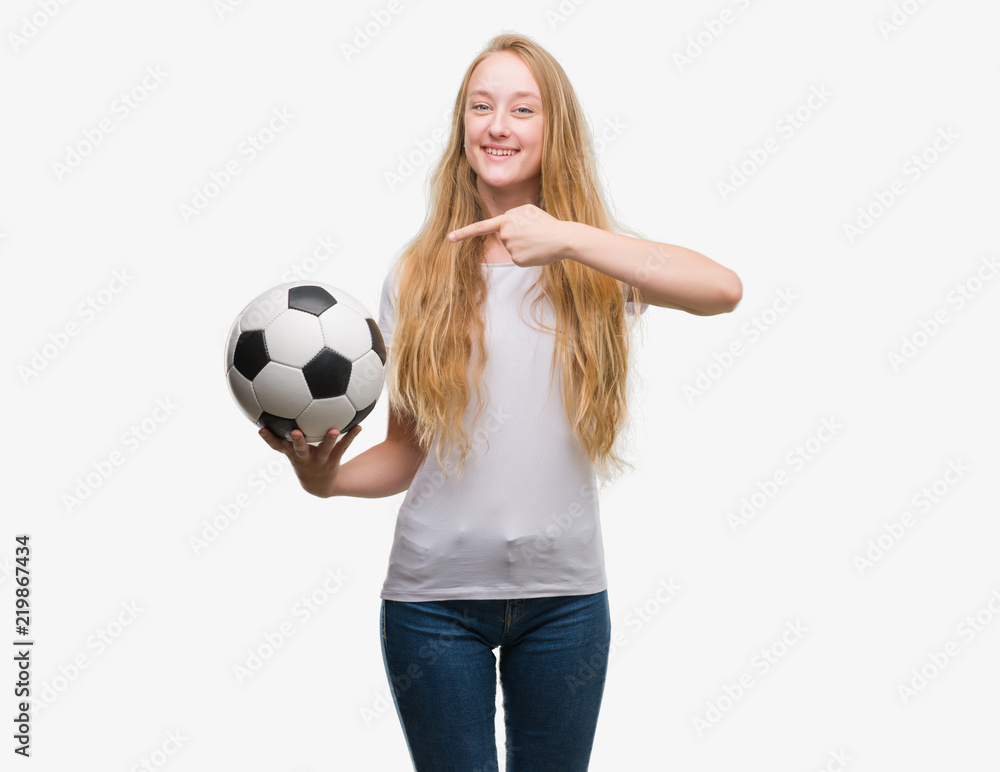 Blonde teenager woman holding soccer football ball very happy pointing with hand and finger