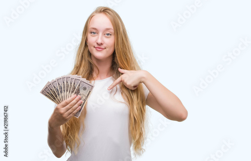Blonde teenager woman holding dollars with surprise face pointing finger to himself