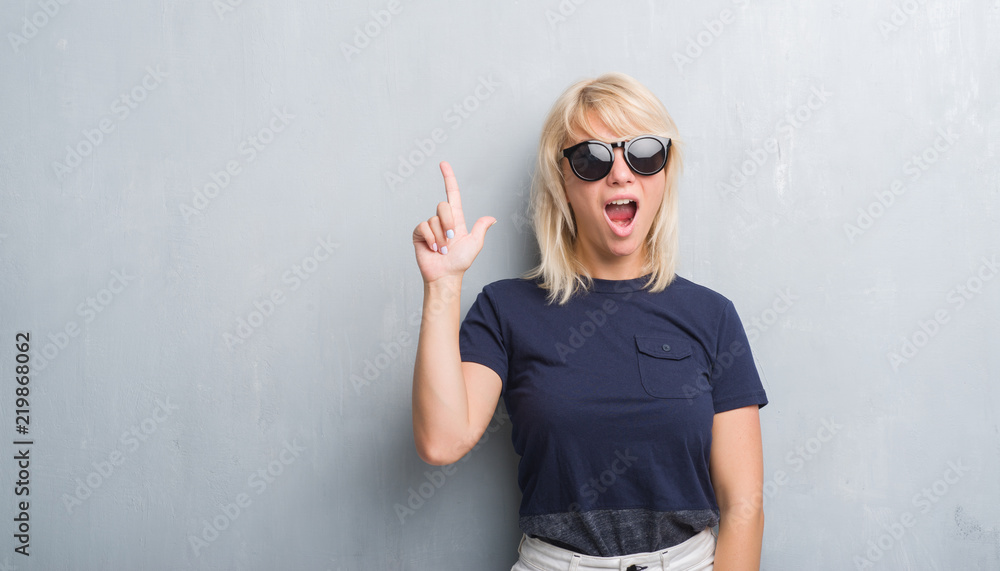 Adult caucasian woman over grunge grey wall wearing sunglasses pointing finger up with successful idea. Exited and happy. Number one. Adult caucasian woman over grunge grey wall wearing sunglasses pointing finger up with successful idea. Exited and happy. Number one.