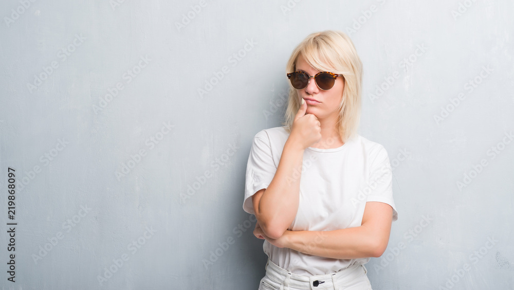 Adult caucasian woman over grunge grey wall wearing sunglasses with hand on chin thinking about question, pensive expression. Smiling with thoughtful face. Doubt concept. Adult caucasian woman over grunge grey wall wearing sunglasses with hand on chin thinking about question, pensive expression. Smiling with thoughtful face. Doubt concept.