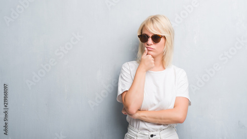 Adult caucasian woman over grunge grey wall wearing sunglasses with hand on chin thinking about question, pensive expression. Smiling with thoughtful face. Doubt concept.