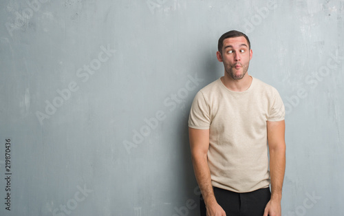 Young caucasian man over grey grunge wall making fish face with lips, crazy and comical gesture. Funny expression.