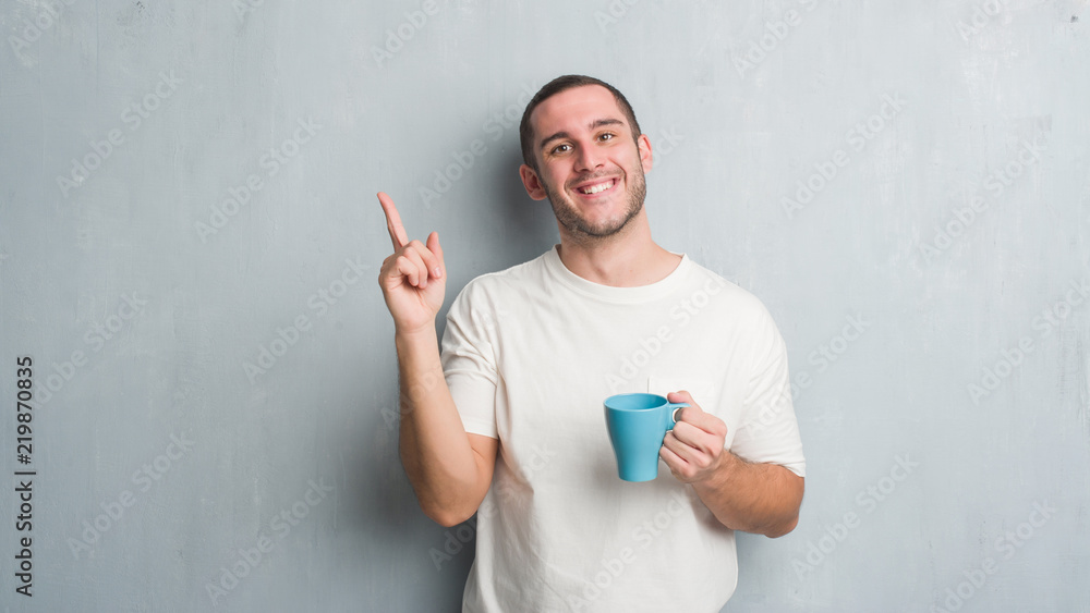Young caucasian man over grey grunge wall driking a cup of coffee very happy pointing with hand and finger to the side Young caucasian man over grey grunge wall driking a cup of coffee very happy pointing with hand and finger to the side
