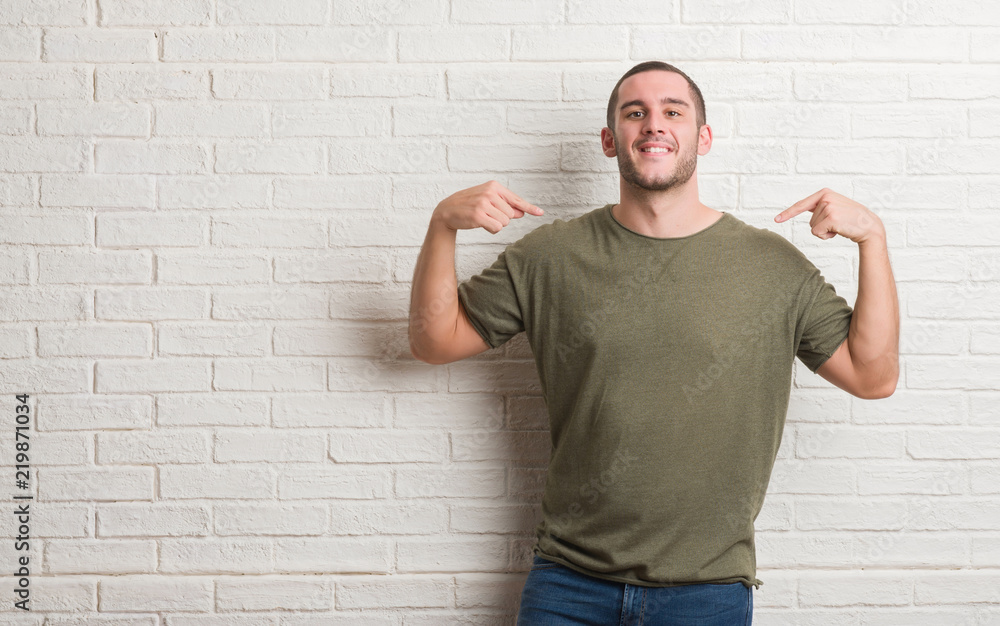 Young caucasian man standing over white brick wall looking confident with smile on face, pointing oneself with fingers proud and happy. Young caucasian man standing over white brick wall looking confident with smile on face, pointing oneself with fingers proud and happy.