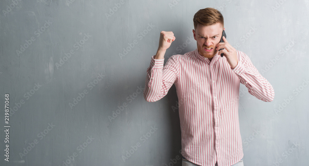 Young redhead man over grey grunge wall talking on the phone annoyed and frustrated shouting with anger, crazy and yelling with raised hand, anger concept