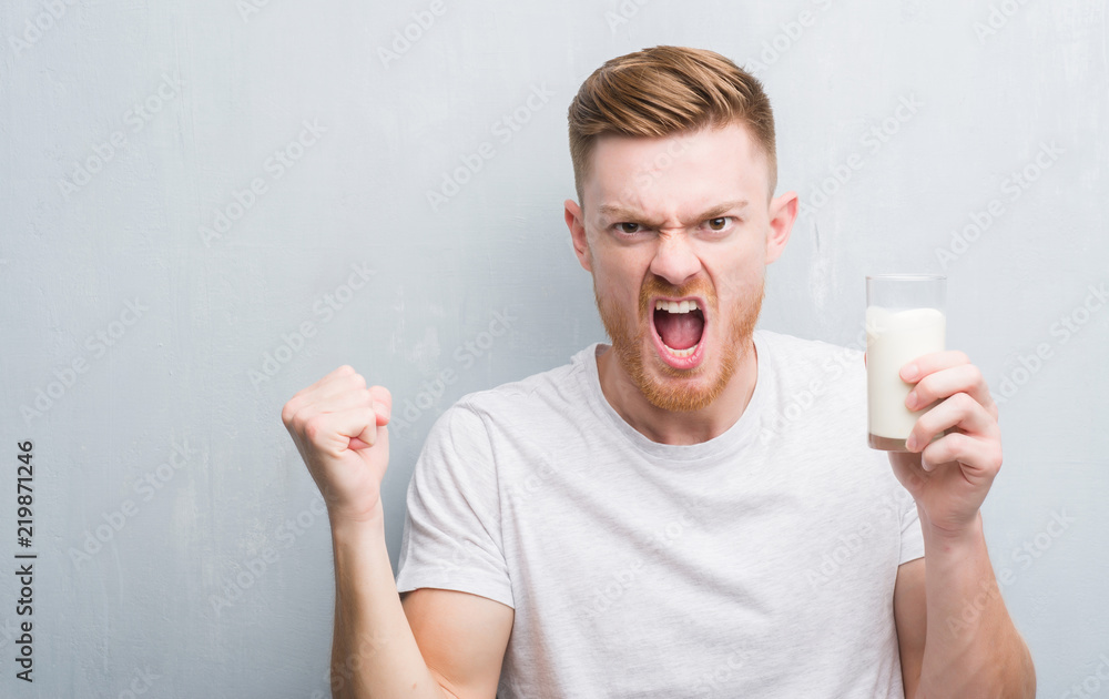 Young redhead man over grey grunge wall drinking a glass of milk annoyed and frustrated shouting with anger, crazy and yelling with raised hand, anger concept Young redhead man over grey grunge wall drinking a glass of milk annoyed and frustrated shouting with anger, crazy and yelling with raised hand, anger concept