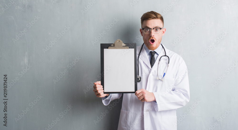 Young redhead doctor man over grey grunge wall holding clipboard scared in shock with a surprise face, afraid and excited with fear expression Young redhead doctor man over grey grunge wall holding clipboard scared in shock with a surprise face, afraid and excited with fear expression