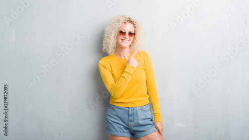 Young blonde woman with curly hair over grunge grey background cheerful with a smile of face pointing with hand and finger up to the side with happy and natural expression on face