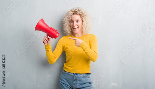 Young blonde woman over grunge grey background holding megaphone very happy pointing with hand and finger