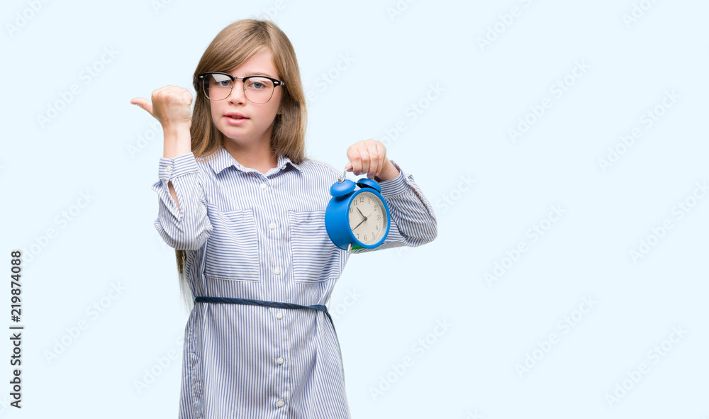 Young blonde child holding alarm clock pointing with hand and finger up with happy face smiling Young blonde child holding alarm clock pointing with hand and finger up with happy face smiling