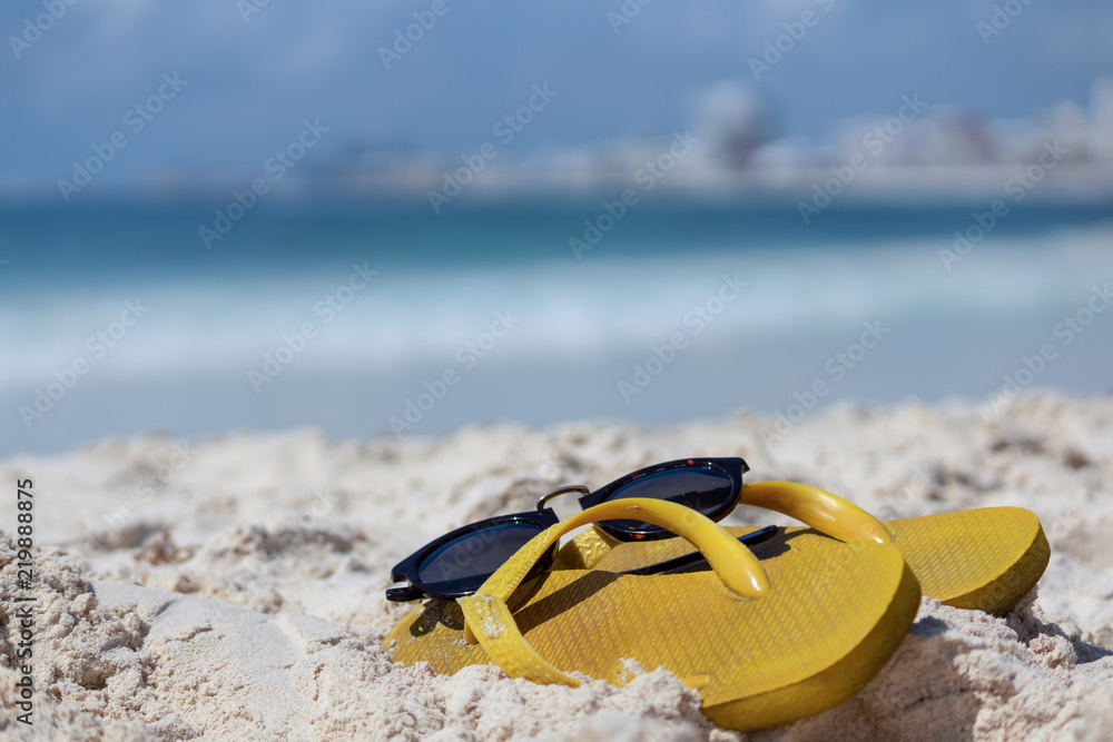 Obraz premium summer snapshot. Flip-Flops and sunglasses in the sand. Focus on sandal details. Caribbean sea in the background.