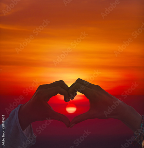 Silhouette of couple making heart - shape symbol with sunset above ocean   sea horizon. Sun is in small natural out of focus.