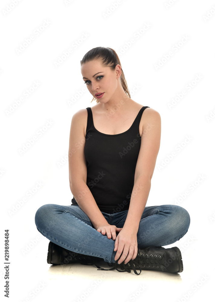full length portrait of brunette girl wearing black single and jeans. seated pose. isolated on white studio background.
