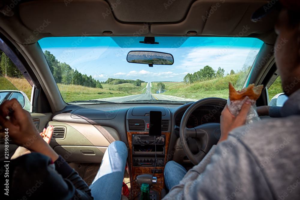 Naklejka premium Guy and woman inside the car driving and eat pie on the country roadway between fields with brown grass and snowy mountains . Sun is shining. Shoot from the back