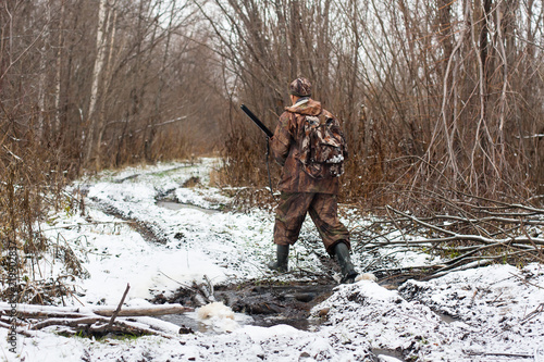 hunter with hunting gun crosses stream in winter